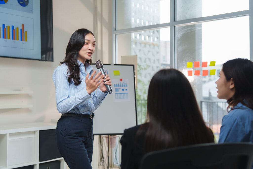 A young professional holding a microphone and giving a presentation with charts and graphs to coworkers in an office meeting.
